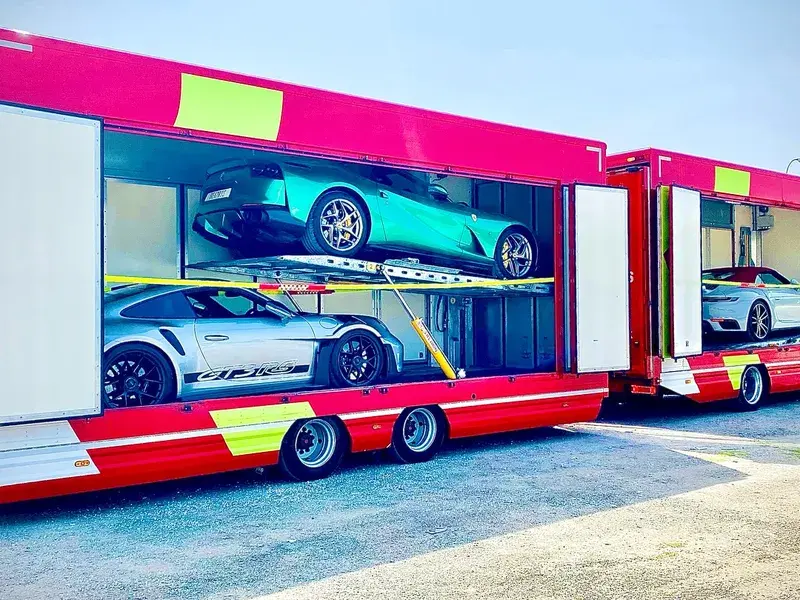 Two red transport trucks with transparent sides, each carrying two luxury sports cars, parked on a sunny day.